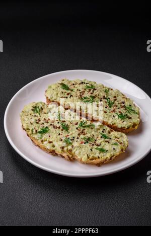 Toast grillé avec pâte d'avocat, œufs, fromage à la crème, sel, épices et herbes sur une table en béton texturé Banque D'Images