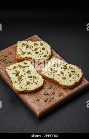 Toast grillé avec pâte d'avocat, œufs, fromage à la crème, sel, épices et herbes sur une table en béton texturé Banque D'Images