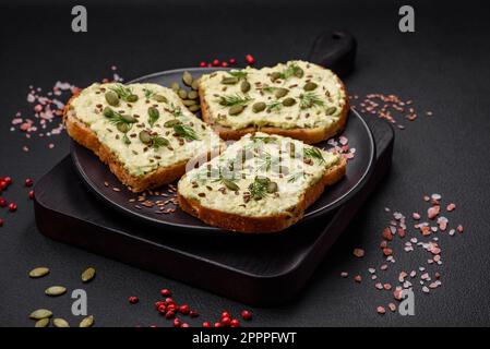 Toast grillé avec pâte d'avocat, œufs, fromage à la crème, sel, épices et herbes sur une table en béton texturé Banque D'Images