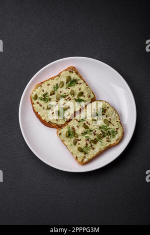 Toast grillé avec pâte d'avocat, œufs, fromage à la crème, sel, épices et herbes sur une table en béton texturé Banque D'Images