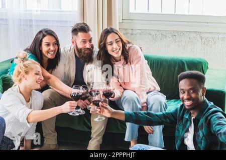 Groupe d'amis pour un apéritif à la maison avec vin rouge assis sur le canapé. Les personnes multiethniques du millénaire apprécient la fête qui se réunit. La vie Banque D'Images