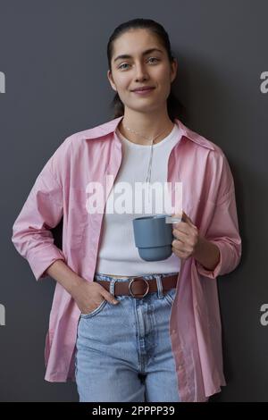 Image verticale d'une jeune femme souriant à l'appareil photo debout avec une tasse de café sur fond gris Banque D'Images
