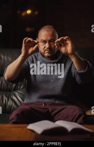 Un homme d'âge moyen tient des lunettes en écrevisses dans une pièce sombre. Problèmes de vision, lunettes de lecture. Banque D'Images