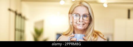 Belle femme d'affaires mûre dans des verres souriant à l'appareil photo et à boire café tout en travaillant sur l'ordinateur portable dans le bureau Banque D'Images