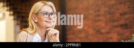 Femme d'affaires gaie et mûre dans des verres souriant, utilisant un ordinateur portable et buvant du café tout en travaillant au bureau Banque D'Images