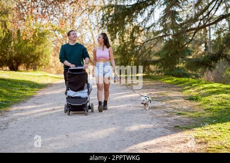 Un jeune couple fait une promenade tranquille dans le parc par une belle journée d'été, poussant son bébé dans une poussette et accompagné de son fidèle chien Banque D'Images