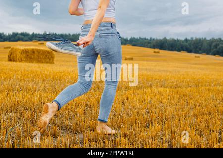 une fille pieds nus avec des baskets à la main marchant dans le champ agricole avec des botte de foin et des balles après la récolte. Banque D'Images