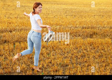 une fille pieds nus avec des baskets à la main marchant dans le champ agricole avec des botte de foin et des balles après la récolte. Banque D'Images