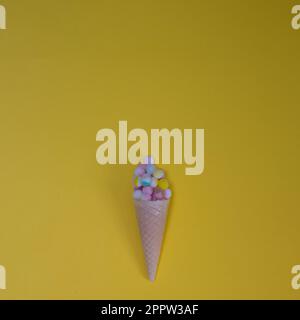 Boules moelleuses de différentes couleurs dans un cône de crème glacée, avec un espace de copie, sur un fond jaune. Scènes de pose plates minimales. Banque D'Images