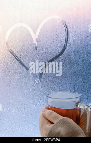 Fermez les mains en buvant du cidre de pomme chaud sous la forme du cœur sur la fenêtre humide Banque D'Images