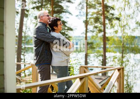 Couple d'âge mûr debout sur le porche et embrassant. Ils sont souriants et regardent la vue magnifique - la forêt et le lac. Un couple senior heureux s'y embrasse Banque D'Images