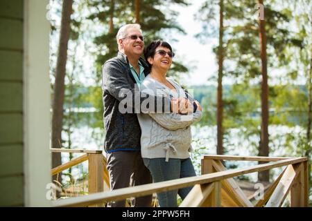 Couple d'âge mûr debout sur le porche et embrassant. Ils sont souriants et regardent la vue magnifique - la forêt et le lac. Un couple senior heureux s'y embrasse Banque D'Images