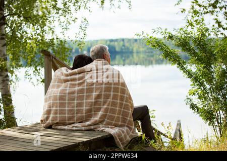 Couple d'âge mûr assis sur des marches en bois et embrassant. Ils sont enveloppés dans une couverture, souriant et regardant la vue magnifique - forêt et lac. Joyeux senior Banque D'Images