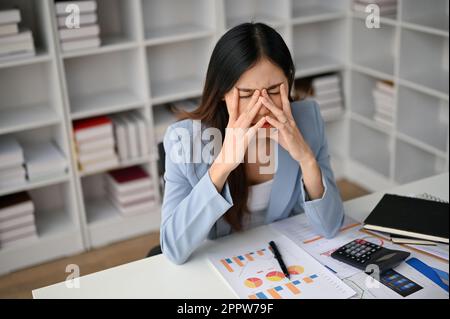 Une femme d'affaires asiatique millénaire fatiguée et stressée s'assoit à son bureau, couvrant son visage et ses pleurs, souffrant de tension oculaire. Banque D'Images