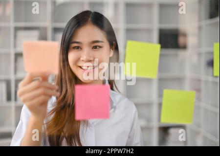 Une employée de bureau ou une employée de marketing asiatique créative et inspirée du millénaire lisant des notes adhésives sur un mur de verre de son bureau. Banque D'Images