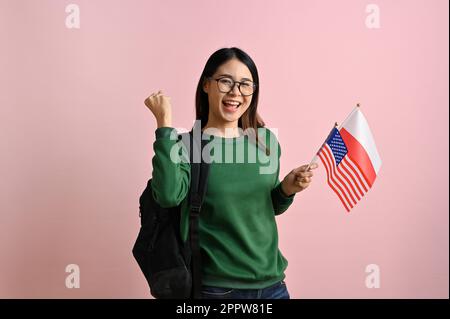 Jolie jeune asiatique étudiante à l'université tenant les drapeaux de la Pologne et des États-Unis et montrant le geste Oui, célébrant et réjouissant, se tient isolé sur un Banque D'Images