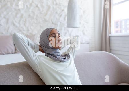 Jeune femme musulmane se reposant à la maison avec les yeux fermés les mains derrière la tête se nouant assis sur le canapé, femme dans le hijab de près dans le salon. Banque D'Images