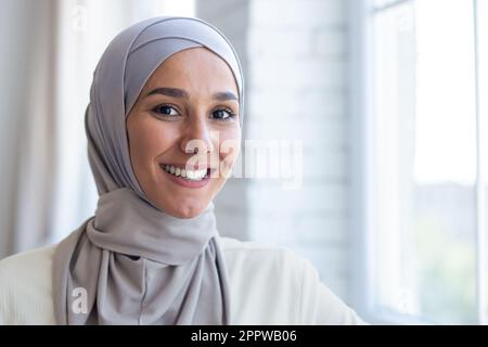 Portrait en gros plan de la jeune belle femme arabe, femme musulmane en hijab souriant et regardant l'appareil photo, debout près de la fenêtre à la maison. Banque D'Images