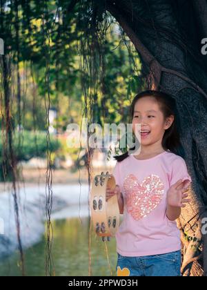 Petite fille asiatique jouer le tambourin, dans le jardin sous le grand arbre Banque D'Images