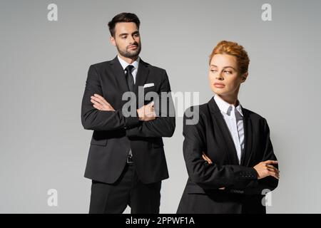 homme d'affaires sceptique en costume élégant regardant la femme rouge dans l'habillement formel isolé sur gris, image de stock Banque D'Images