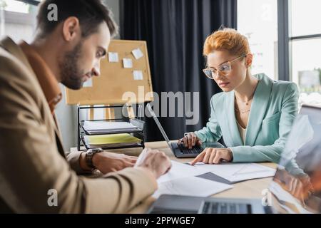 redhead femme d'affaires en lunettes de vue en utilisant un ordinateur portable et en pointant vers des papiers près d'un collègue flou dans le bureau, image de stock Banque D'Images