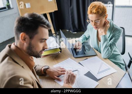 homme d'affaires barbu pointant vers des infographies près d'une femme dans des lunettes travaillant sur un ordinateur portable au bureau, image de stock Banque D'Images