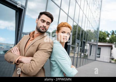 les hommes d'affaires sceptiques se tenant dos à dos avec les bras pliés près du bâtiment sur la rue de la ville, image de stock Banque D'Images
