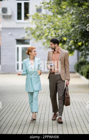 pleine longueur de femme d'affaires joyeuse qui parle à un collègue tout en marchant dans la rue urbaine, image de stock Banque D'Images