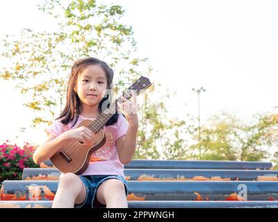 Petite fille asiatique jouer l'ukulele, dans le jardin sur le tuyau en acier Banque D'Images