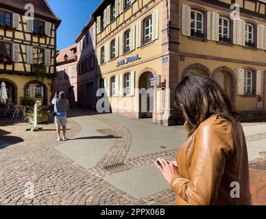 Ribeauville, France, 22 septembre 2022 : une femme envoie des textes sur son smartphone, en admirant la vue urbaine d'une place publique avec la banque du crédit mutuel et l'hôtel de V. Banque D'Images