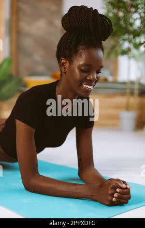 Femme afro-américaine durable avec des dreadlocks debout dans le coude de la planche à travers la douleur de gros plan, l'entraînement sur le tapis de gymnastique dans le studio décoré. Muscle Banque D'Images