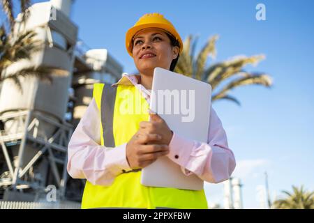 Faible angle de jeune femme ethnique africaine américaine constructeur en uniforme et casque debout tenant un ordinateur portable tout en regardant le projet pendant le travail Banque D'Images