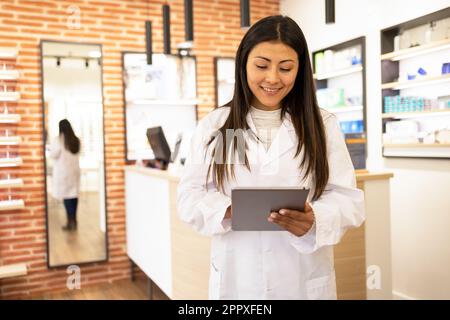 Jeune opticienne souriante en uniforme blanc utilisant la table numérique regardant l'écran dans la boutique optique Banque D'Images