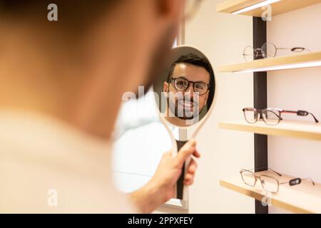 Un jeune client de sexe masculin choisit des lunettes tout en souriant et en regardant le miroir pendant sa visite dans un magasin optique Banque D'Images
