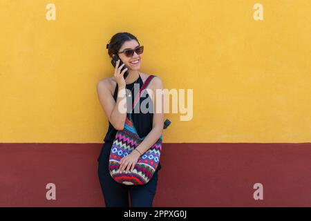 Jeune femme positive dans une tenue tendance et des lunettes de soleil souriant et regardant loin tout en se tenant contre le mur jaune et parlant sur le smartphone à l'extérieur dans Banque D'Images