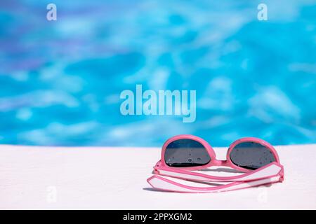 Lunettes de soleil rose vif avec gouttes d'eau placées sur la surface blanche contre la piscine arrière-plan flou Banque D'Images