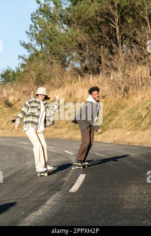 Pleine longueur de jeune couple heureux dans des vêtements décontractés à la planche à roulettes sur la route d'asphalte entre les arbres verts et les buissons pendant la journée Banque D'Images