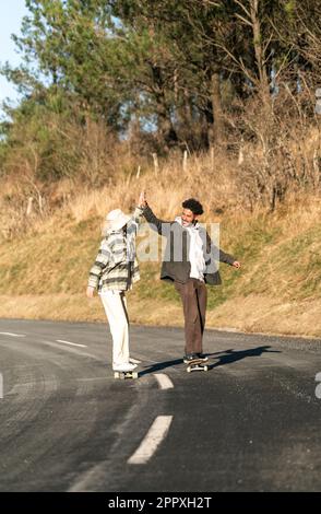 Toute la longueur de jeune couple heureux en vêtements décontractés tenant les mains tout en faisant du skateboard sur la route asphaltée entre les arbres verts et les buissons pendant la journée Banque D'Images