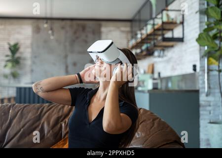 Jeune femme dans des lunettes VR jouant à un jeu vidéo tout en étant assise sur un canapé dans le salon Banque D'Images