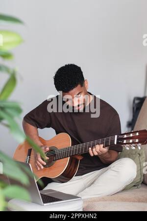 Un jeune musicien afro-américain d'origine ethnique positif dans une tenue décontractée regardant un tutoriel vidéo sur ordinateur portable tout en jouant de la guitare acoustique sur les confortables s. Banque D'Images