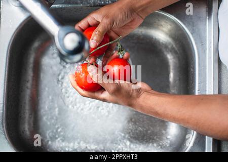 Grand angle de l'anonyme africain américain chef laver des tomates rouges mûres fraîches dans évier chromé avec eau du robinet tout en préparant des ingrédients pour la cuisine Banque D'Images