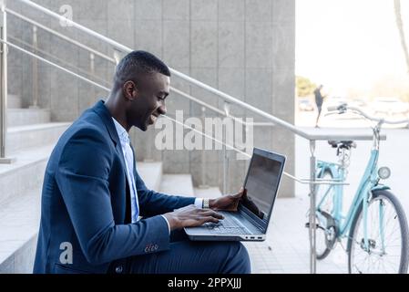 Vue latérale d'un homme d'affaires afro-américain souriant vêque de vêtements habillés, assis dans les escaliers et dactylographiant sur un ordinateur portable, tout en travaillant à distance près d'un homme de la ville Banque D'Images