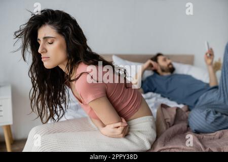 Une femme brunette déçue se sent mal lorsqu'elle est assise près d'un petit ami flou avec un smartphone sur le lit à la maison, image de stock Banque D'Images