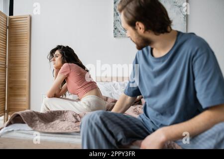 Femme indifférente dans un pyjama assis sur le lit près d'un petit ami flou à la maison, image de stock Banque D'Images
