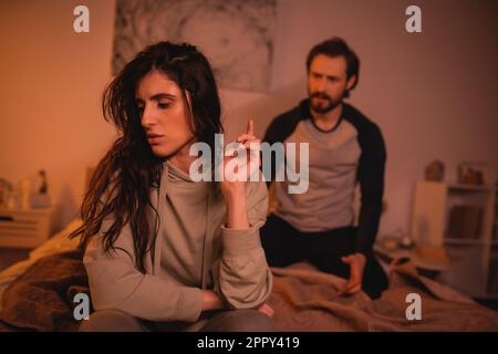 Triste femme brune assise près d'un petit ami flou sur le lit la nuit, image de stock Banque D'Images