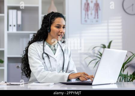 Femme hispanique médecin travaillant à l'intérieur du bureau médical à un bureau, femme tapant sur un ordinateur portable, souriant avec un casque d'appel vidéo, femme médecin consultant des patients à distance en ligne. Banque D'Images