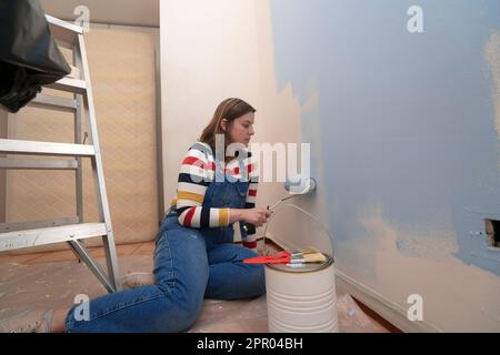 Femme agenouillée vêtue de combinaisons et blouse rayée, vue de profil, peinture d'un mur blanc avec un rouleau de peinture bleue, à l'intérieur d'une chambre vide avec un Banque D'Images