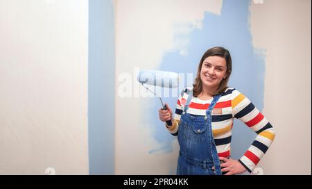 Belle femme debout vêtue de combinaisons, souriant à l'appareil photo avec rouleau de peinture bleu dans sa main sur fond de mur blanc à moitié peint Banque D'Images