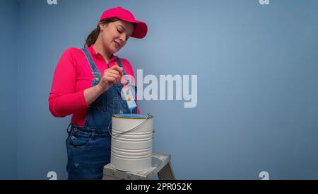 Belle femme vêtue de combinaisons et t-shirt rouge debout sur une échelle en métal prenant la brosse hors du pot de peinture bleue à côté d'un mur bleu Banque D'Images