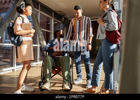 Étudiant handicapé assis sur un fauteuil roulant et parlant à des camarades de classe pendant qu'ils se tiennent dans le couloir de l'école pendant la pause Banque D'Images